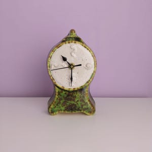 Może przedstawiać: Zielony i brązowy zegar ceramiczny z białą tarczą i czarnymi wskazówkami. Zegar ma kształt dzwonu i posiada unikalny design.