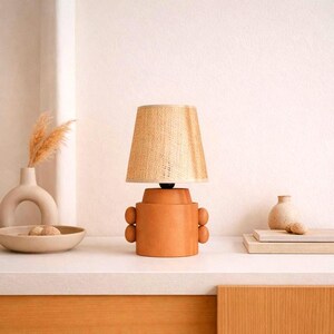 A terracotta-colored table lamp with a woven beige lampshade. The lamp has a cylindrical base with rounded accents- buttons. The lamp is on a white surface, with decorative vases and a bowl in the background.