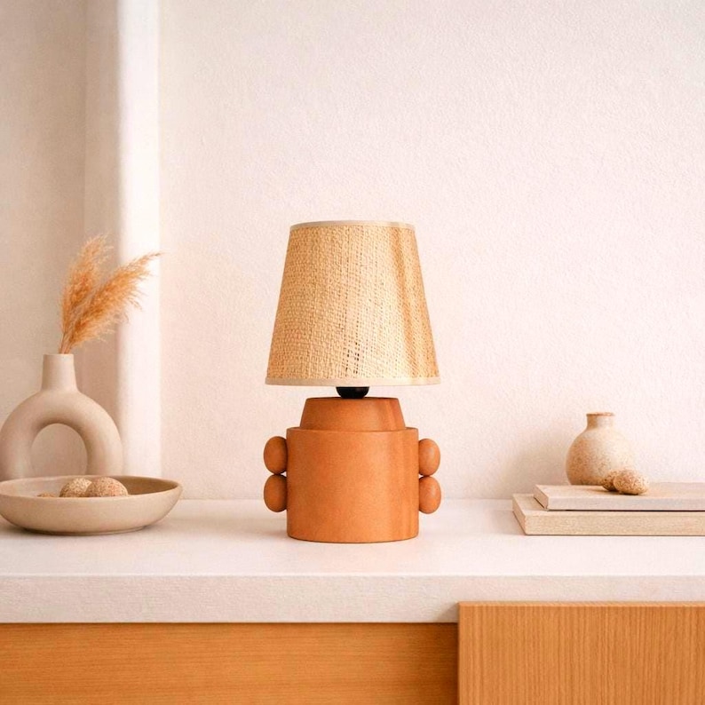 A terracotta-colored table lamp with a woven beige lampshade. The lamp has a cylindrical base with rounded accents- buttons. The lamp is on a white surface, with decorative vases and a bowl in the background.