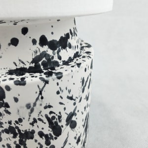 May include: White ceramic vase with black splatter paint design. The vase has a rounded base and a cylindrical top.