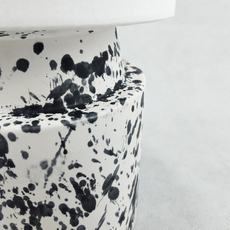 May include: White ceramic vase with black splatter paint design. The vase has a rounded base and a cylindrical top.