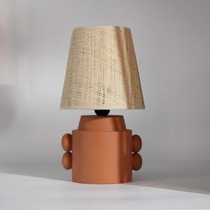 May include: A terracotta table lamp with a woven beige lampshade. The lamp base has a cylindrical shape with rounded accents on the sides. The lampshade is conical.
