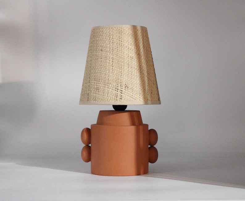 A terracotta-colored table lamp with a woven beige lampshade. The lamp base has a cylindrical shape with rounded accents on the sides. The lampshade is conical.