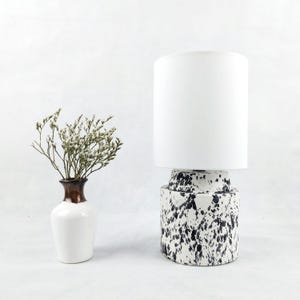 May include: A white ceramic table lamp with a white fabric shade. The base of the lamp has a black and white splatter pattern.