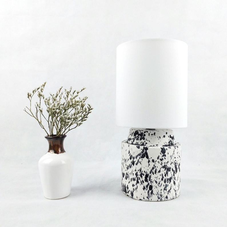 May include: A white ceramic table lamp with a white fabric shade. The base of the lamp has a black and white splatter pattern.