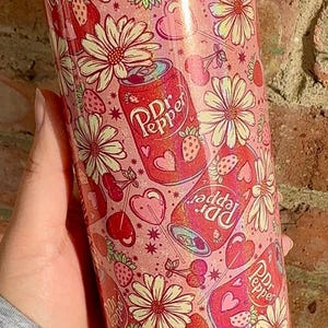 May include: A pink and white tumbler with a repeating pattern of Dr Pepper cans, daisies, strawberries, cherries, and hearts. The tumbler has a clear lid.