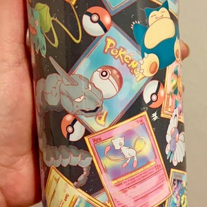 May include: A black and white tumbler with a colorful pattern of Pokemon cards and Pokeballs. The tumbler has a clear lid.