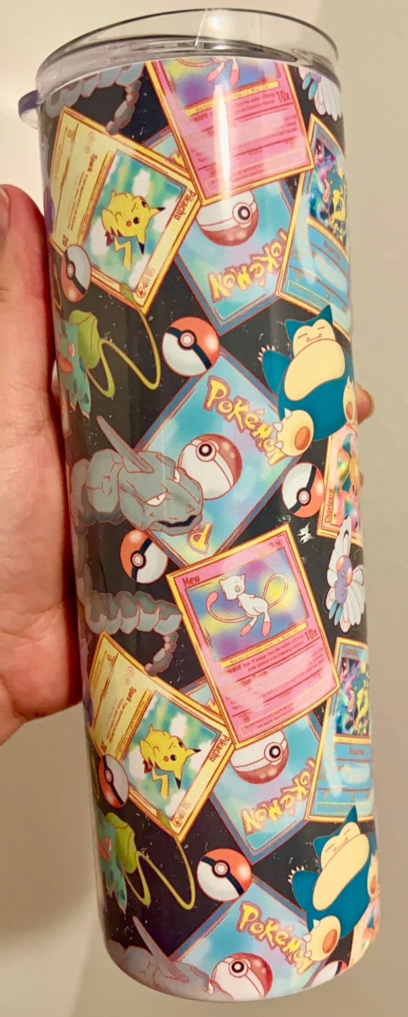 May include: A black and white tumbler with a colorful pattern of Pokemon cards and Pokeballs. The tumbler has a clear lid.