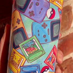 May include: A blue and green tumbler with a pattern of Pokemon game cartridges and Game Boy consoles. The tumbler has a clear lid.