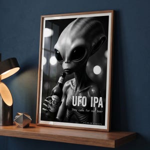 Op de afbeelding: Zwart-wit ingelijste print met een grijze alien die een bierfles vasthoudt. De print leest "UFO IPA" met de slogan "They came for our Beer." De alien heeft grote zwarte ogen en een slank postuur.