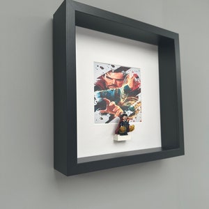 Può includere: Opera d'arte incorniciata con un'immagine colorata di Doctor Strange con una piccola statuina del personaggio attaccata. La cornice nera è montata su un muro grigio. L'opera d'arte ha un passe-partout bianco.