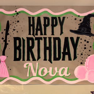 May include: A handmade birthday banner with the words "HAPPY BIRTHDAY Nova" in black and green lettering. The banner features a witch hat, pink balloons, a pink tassel, and star accents, all outlined in pink and green.