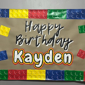 May include: A brown paper banner with a Lego theme. The banner says "Happy Birthday Kayden" in black and white lettering. The banner is decorated with colorful Lego blocks.
