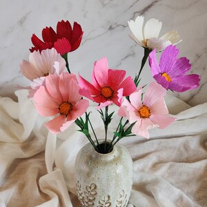7 cosmos, red, white, lilac, light lilac, pink, light pink, and hot pink are in a texted vase against a marble background on a table covered with white cotton.