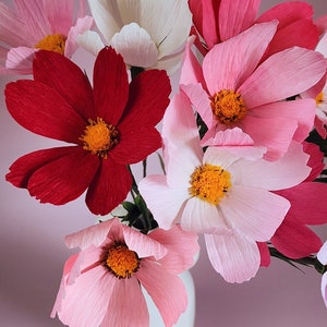 May include: A bouquet of paper flowers in a white vase. The flowers are pink, red, and white and have a delicate, layered texture.