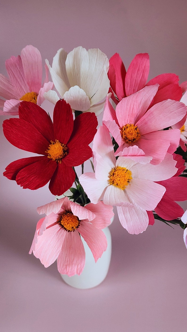 May include: A bouquet of paper flowers in a white vase. The flowers are pink, red, and white and have a delicate, layered texture.