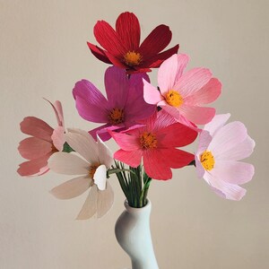 May include: A bouquet of pink, red, and white paper flowers in a light blue ceramic vase. The vase has a unique, curvy shape.