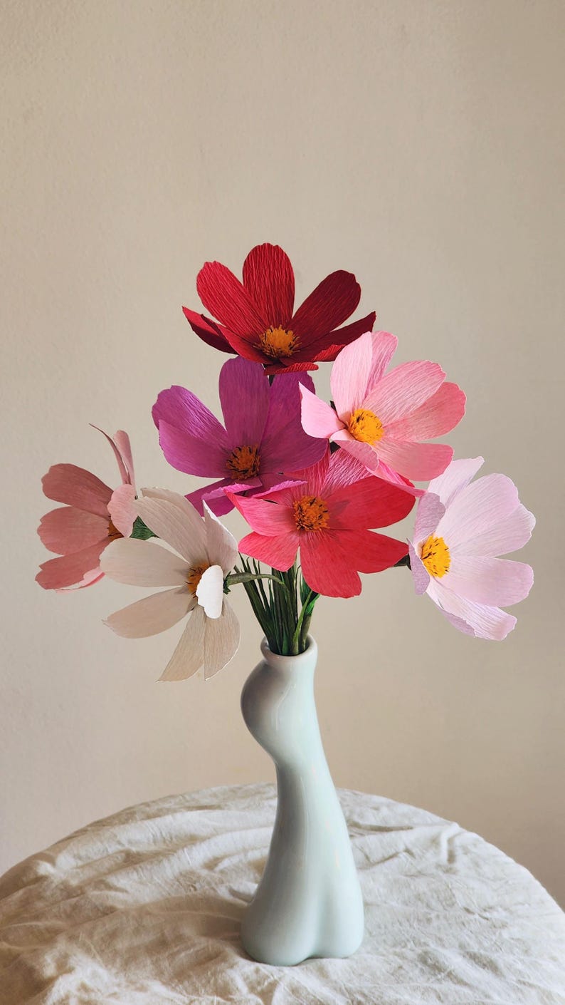 May include: A bouquet of pink, red, and white paper flowers in a light blue ceramic vase. The vase has a unique, curvy shape.