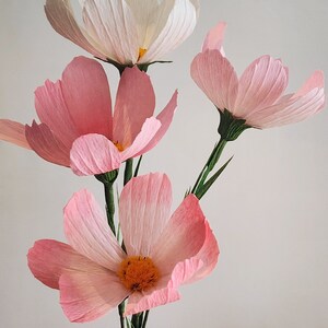 May include: A bouquet of four paper flowers, three pink and one white, with green stems. The flowers have a delicate, layered texture.