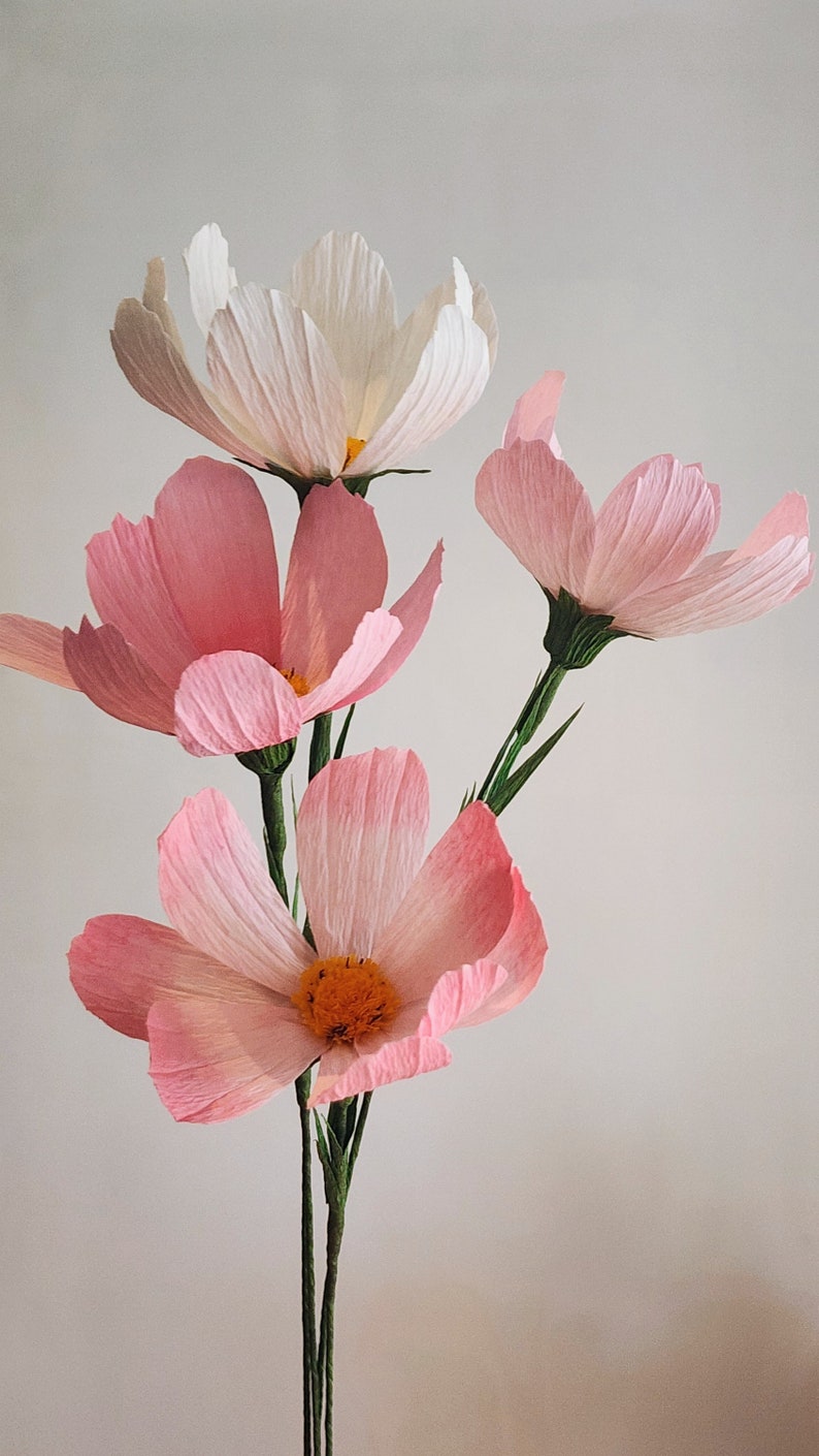 May include: A bouquet of four paper flowers, three pink and one white, with green stems. The flowers have a delicate, layered texture.