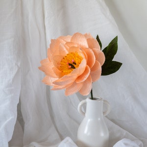 Puede incluir: Una flor de papel de color melocot&oacute;n con un centro amarillo y hojas verdes en un jarr&oacute;n de cer&aacute;mica blanco.