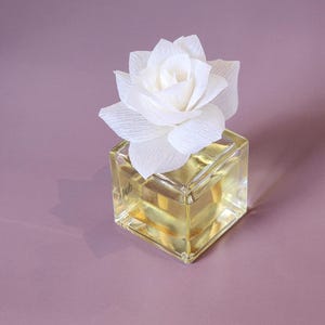 Pode incluir: Um frasco difusor de vidro transparente com uma flor de papel branco no topo. O frasco é quadrado e cheio de um líquido amarelo. A flor é uma única rosa com várias camadas de pétalas.