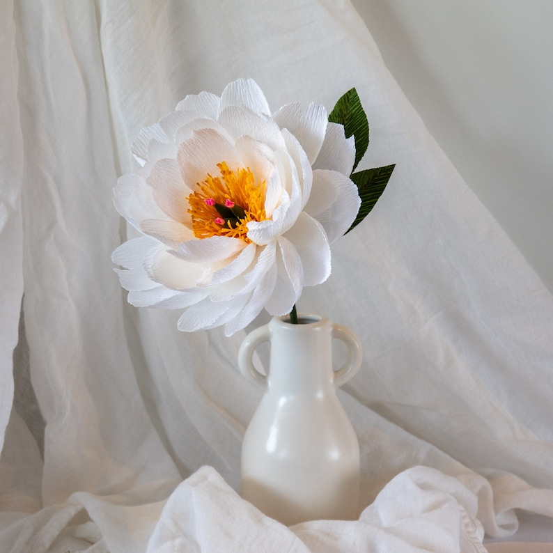 Puede incluir: Una flor de papel blanca con un centro amarillo y detalles rosas en un jarr&oacute;n de cer&aacute;mica blanco. El jarr&oacute;n tiene dos asas y un cuerpo redondeado. La flor y el jarr&oacute;n est&aacute;n sobre un fondo de tela blanca.