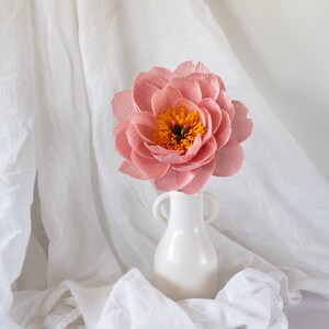 Puede incluir: Una sola flor de papel rosa con un centro amarillo se encuentra en un jarr&oacute;n de cer&aacute;mica blanca. El jarr&oacute;n tiene dos asas. La flor y el jarr&oacute;n est&aacute;n sobre un fondo de tela blanca.
