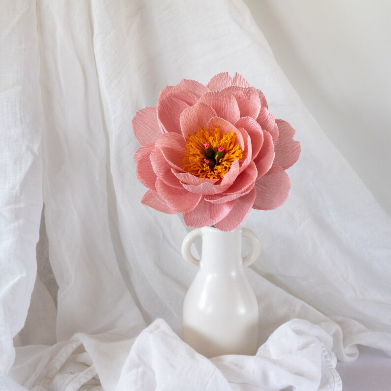 Puede incluir: Una sola flor de papel rosa con un centro amarillo se encuentra en un jarr&oacute;n de cer&aacute;mica blanca. El jarr&oacute;n tiene dos asas. La flor y el jarr&oacute;n est&aacute;n sobre un fondo de tela blanca.
