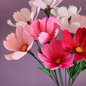 May include: A bouquet of pink and red paper flowers in a white ceramic vase. The flowers are arranged in a vase with a textured surface. The flowers are made of paper and have a delicate, layered appearance.