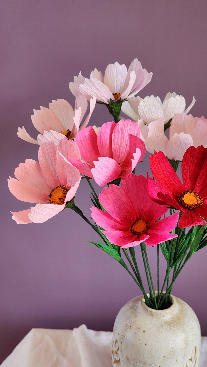 May include: A bouquet of pink and red paper flowers in a white ceramic vase. The flowers are arranged in a vase with a textured surface. The flowers are made of paper and have a delicate, layered appearance.