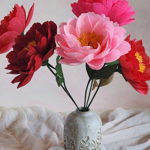 Puede incluir: Un ramo de flores de papel rosa y rojo en un jarr&oacute;n de cer&aacute;mica blanca con un dise&ntilde;o de hojas. Las flores est&aacute;n dispuestas en un jarr&oacute;n sobre un mantel de lino blanco.