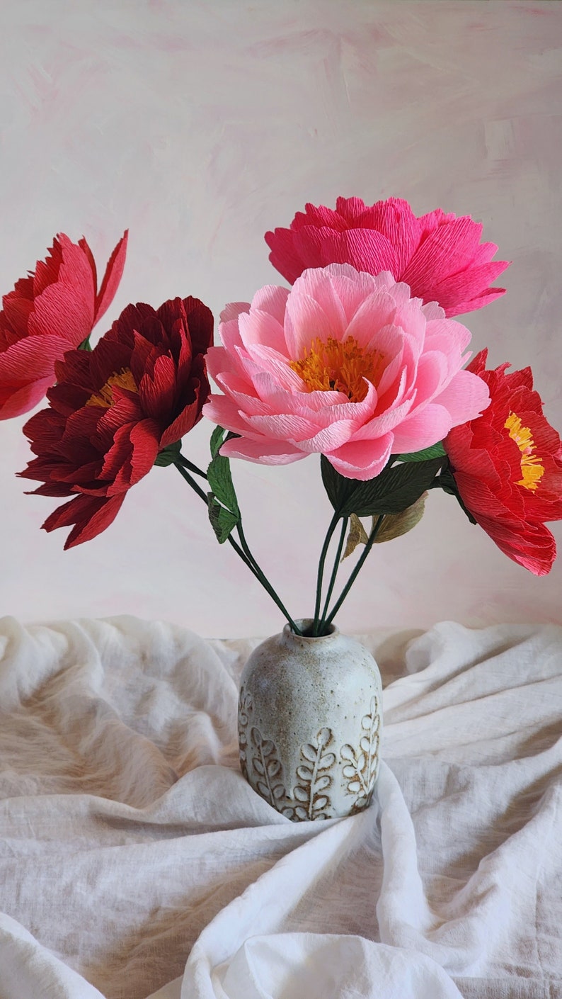 Puede incluir: Un ramo de flores de papel rosa y rojo en un jarr&oacute;n de cer&aacute;mica blanca con un dise&ntilde;o de hojas. Las flores est&aacute;n dispuestas en un jarr&oacute;n sobre un mantel de lino blanco.