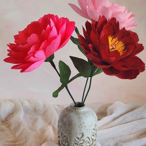 Puede incluir: Tres grandes flores de papel en un jarr&oacute;n de cer&aacute;mica blanco con un dise&ntilde;o de hojas. Las flores son rosas, rojas y de un color fucsia brillante.