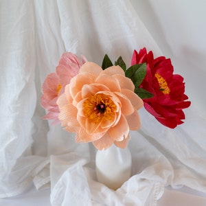 Puede incluir: Un ramo de tres flores de papel en un jarr&oacute;n blanco. Las flores son de color melocot&oacute;n, rosa y rojo. Las flores est&aacute;n dispuestas en un estilo suelto y natural.