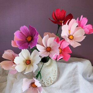 May include: A bouquet of pink, white, and red paper flowers in a white ceramic vase with a leaf design. The flowers are arranged in a vase on a white tablecloth.
