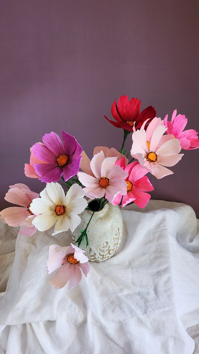 May include: A bouquet of pink, white, and red paper flowers in a white ceramic vase with a leaf design. The flowers are arranged in a vase on a white tablecloth.