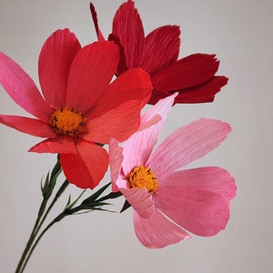 May include: Three paper flowers in shades of pink and red. The flowers are made of crinkled paper and have a delicate, feminine look.