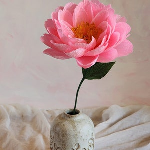 Puede incluir: Una sola flor de papel rosa con un centro amarillo en un jarr&oacute;n de cer&aacute;mica blanco con un dise&ntilde;o de hojas. El jarr&oacute;n est&aacute; sobre una superficie de tela blanca.