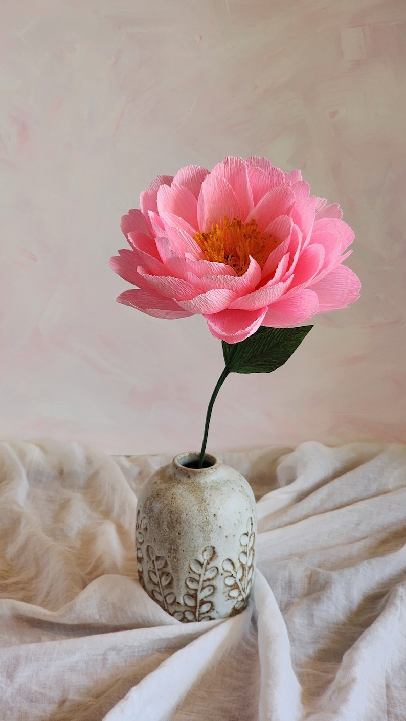 Puede incluir: Una sola flor de papel rosa con un centro amarillo en un jarr&oacute;n de cer&aacute;mica blanco con un dise&ntilde;o de hojas. El jarr&oacute;n est&aacute; sobre una superficie de tela blanca.