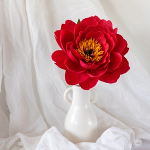 Puede incluir: Una sola flor de peon&iacute;a de papel roja en un jarr&oacute;n de cer&aacute;mica blanco. La flor est&aacute; en plena floraci&oacute;n y tiene un centro amarillo con acentos rojos.