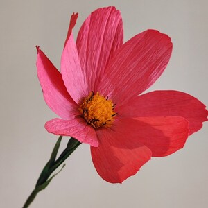 May include: A single pink paper flower with a yellow center. The flower is on a stem with green leaves.