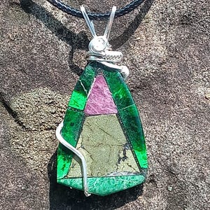 May include: A pendant featuring a triangular mosaic of green, pink, and metallic gold-toned stone segments held by a silver-colored wire wrap. The pendant is suspended from a black cord necklace, showcasing a geometric arrangement of colored minerals.