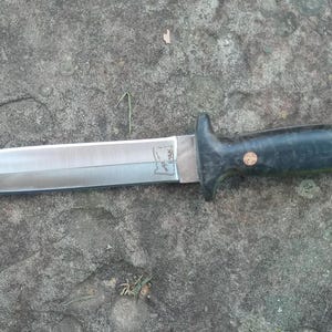 Puede incluir: Una daga de doble filo con una hoja de acero pulido y un mango oscuro y texturizado. El mango está asegurado con dos remaches de cobre. La daga descansa sobre una superficie de piedra gris rugosa.