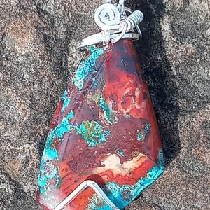 May include: A handcrafted pendant featuring a vibrant, irregularly shaped stone with a mix of red, turquoise, and cream colors. The stone is encased in a silver wire setting and hangs from a black cord. The pendant's unique pattern and color palette make it a striking piece of jewelry.
