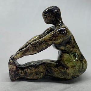 May include: A ceramic sculpture of a person in a seated position with their legs stretched out in front of them. The sculpture is painted in shades of green and black.