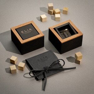 以下が含まれることがあります： 木製の蓋が付いた黒い指輪の箱で、中に金の指輪が入っています。箱は灰色の表面に置かれており、その周りに小さな木製のブロックが散らばっています。また、紐で閉めることができる黒いベルベット製のポーチがあり、そこに「ELIA FINE JEWELRY」という文字が印刷されています。