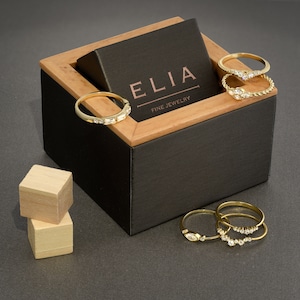 以下が含まれることがあります： 「ELIA FINE JEWELRY」と刻まれた黒い蓋が付いた木製の箱。箱が開いており、ダイヤモンドがついた金の指輪がいくつか入っています。指輪は積み重ねられています。