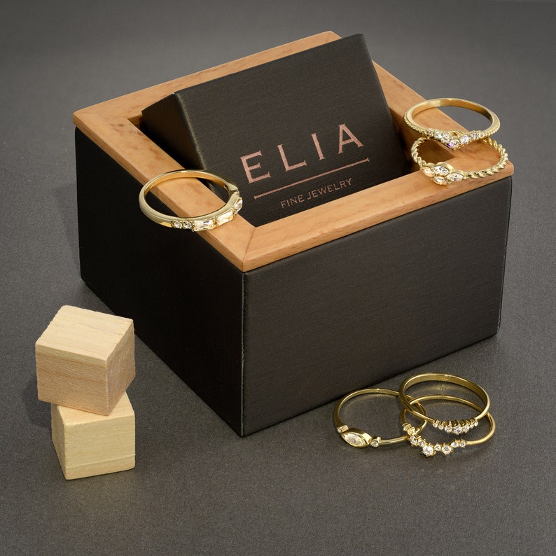 以下が含まれることがあります： 「ELIA FINE JEWELRY」と刻まれた黒い蓋が付いた木製の箱。箱が開いており、ダイヤモンドがついた金の指輪がいくつか入っています。指輪は積み重ねられています。