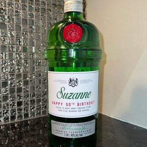 May include: A green glass bottle of Tanqueray gin with a personalized label that reads "Suzanne Happy 50th Birthday Cheers to many more fabulous years from Stan, Brandon and Shavena". The label also includes the text "Charles Tanqueray & Co London 1.14L 40% alc./vol. England".